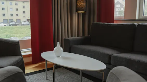 Sofa set surrounds an oval white coffee table with a vase, situated in a living room with red curtains and a floor lamp near the window.