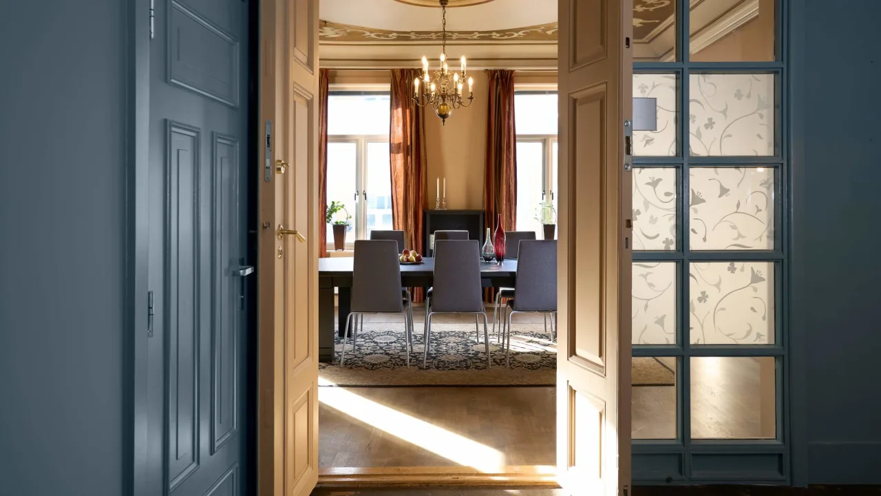 Open doors leading to a dining room with a table, chairs, and a chandelier; sunlight streams in, highlighting the elegant interior.