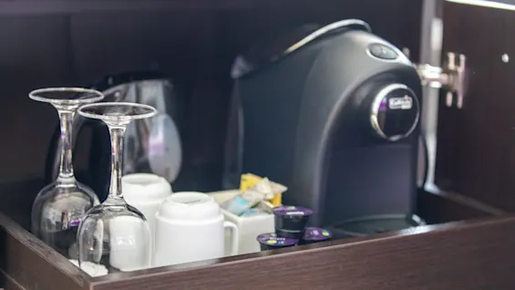 En kaffemaskin står i et treskap ved siden av to vinglass opp ned og kopper, omgitt av kaffekapsler og sukkerpakker.