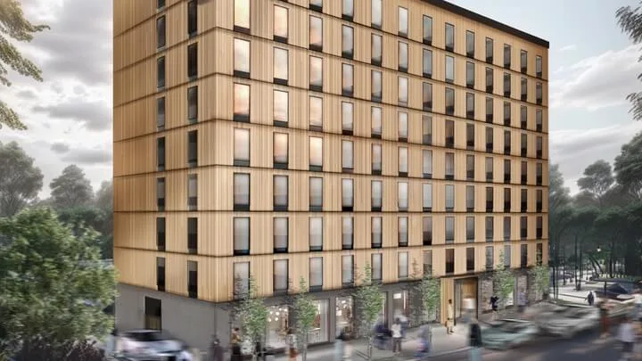 Rakennus seisoo korkeana puujulkisivulla, merkintä "HOTEL." Ihmisiä ja autoja liikkuu puiden reunustamaa katua pitkin. Taivas on pilvinen pehmeän valon siivilöityessä läpi.