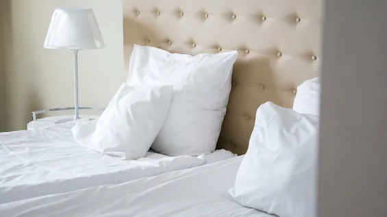 En komfortabel hotelseng med sprøde hvide lagner, bløde puder, en beige tuftet sengegavl og en sengelampe, klar til et afslappende ophold.