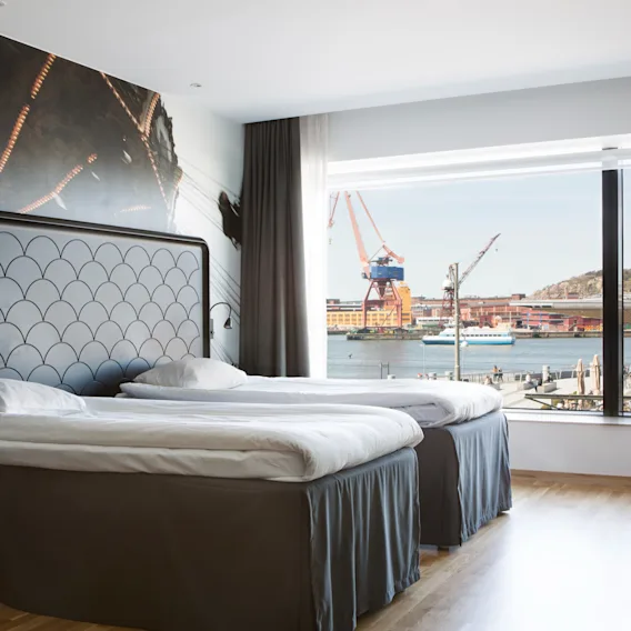 Hotellihuone, jossa kaksi sänkyä ja suuri ikkuna, josta avautuu näkymä vilkkaaseen satamaan nostureineen ja vesi.
