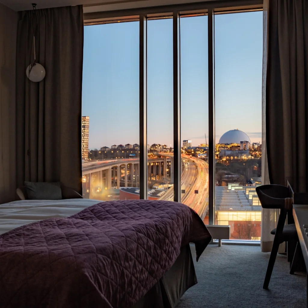 Et hotellrom med en seng med burgunderrødt sengeteppe, mot et stort vindu. Utenfor er et bylandskap synlig i skumringen, inkludert travle veier og en fjern arena.