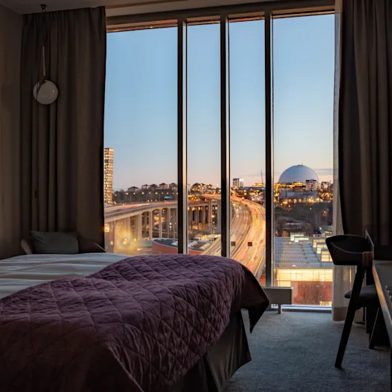 A hotel room features a bed with burgundy bedding, facing a large window. Outside, a cityscape is visible during twilight, including bustling roads and a distant arena.