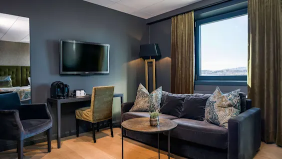 Et moderne opholdsområde med en mørk sofa med mønstrede puder, et rundt sofabord og et vægmonteret TV. Et vindue med tunge gardiner giver udsigt udad.