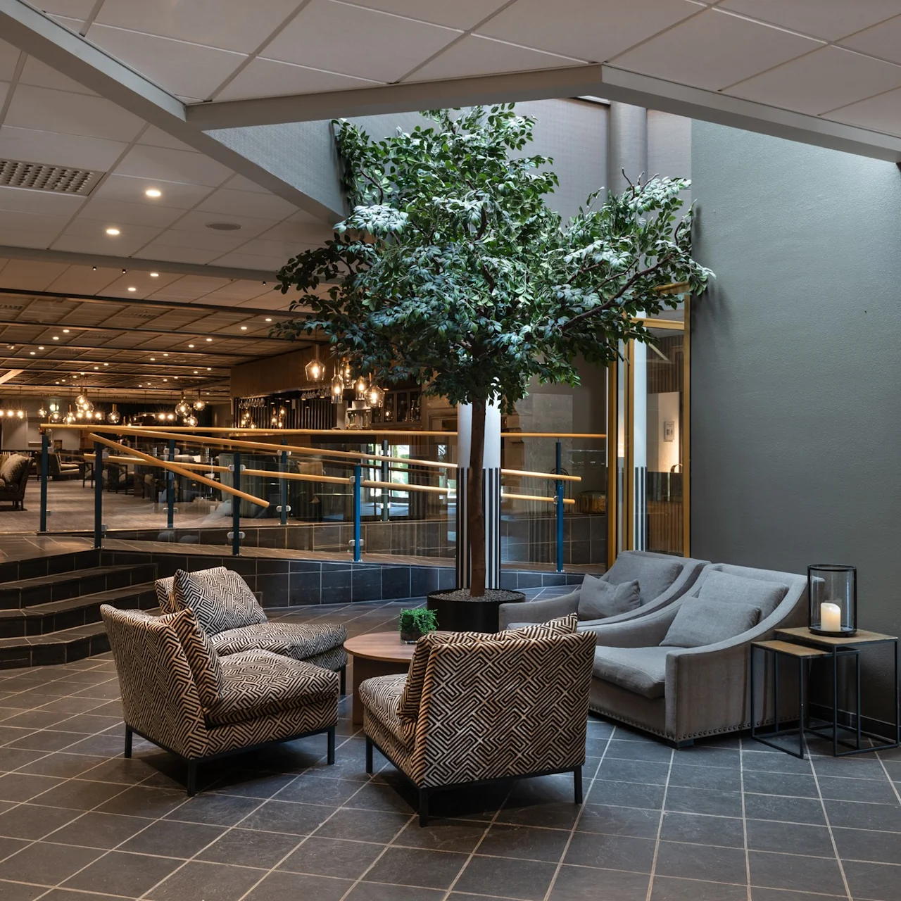 Seating arrangement with patterned armchairs and a small table under a large artificial tree, situated in a modern, well-lit lobby with an elevator on the right.