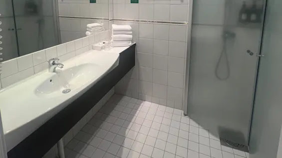 A bathroom features a white-tiled sink with stacked towels beside it, under a large mirror. A glass shower is adjacent, while recessed ceiling lights illuminate the space.