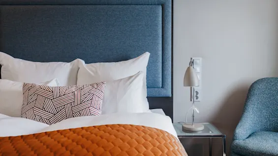 A bed with white pillows and an orange quilt sits beside a nightstand holding a lamp. The setting is a modern bedroom with a blue upholstered chair.