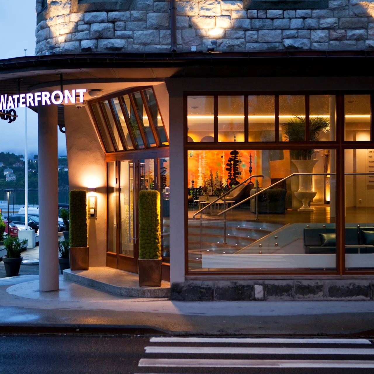 Hotellentré upplyst på natten, glasdörr, upplyst skylt med texten "WATERFRONT", modern inredning synlig. Omgivningen inkluderar parkerade bilar och träd vid vägen.