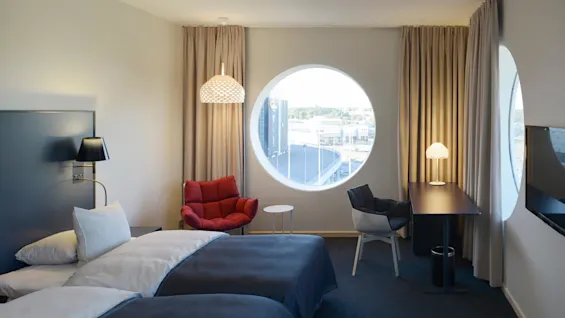 A hotel room features two beds with dark covers, adjacent to a lamp-lit nightstand. A red chair sits near a circular window offering a city view. Desk with chair nearby.