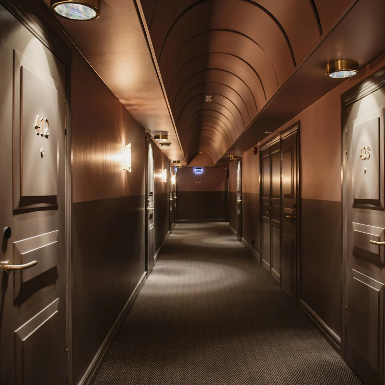 Hotel hallway with numbered doors (412, 413) leading to rooms. Dim lighting casts warm tones. Carpeted floor enhances a cozy atmosphere. Emergency exit sign visible at the corridor's end.