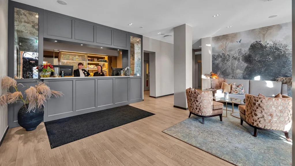 To personer står bak resepsjonsskranken på et hotell, smilende og ivaretar plikter i en moderne dekorert lobby med koselig sitteområde.