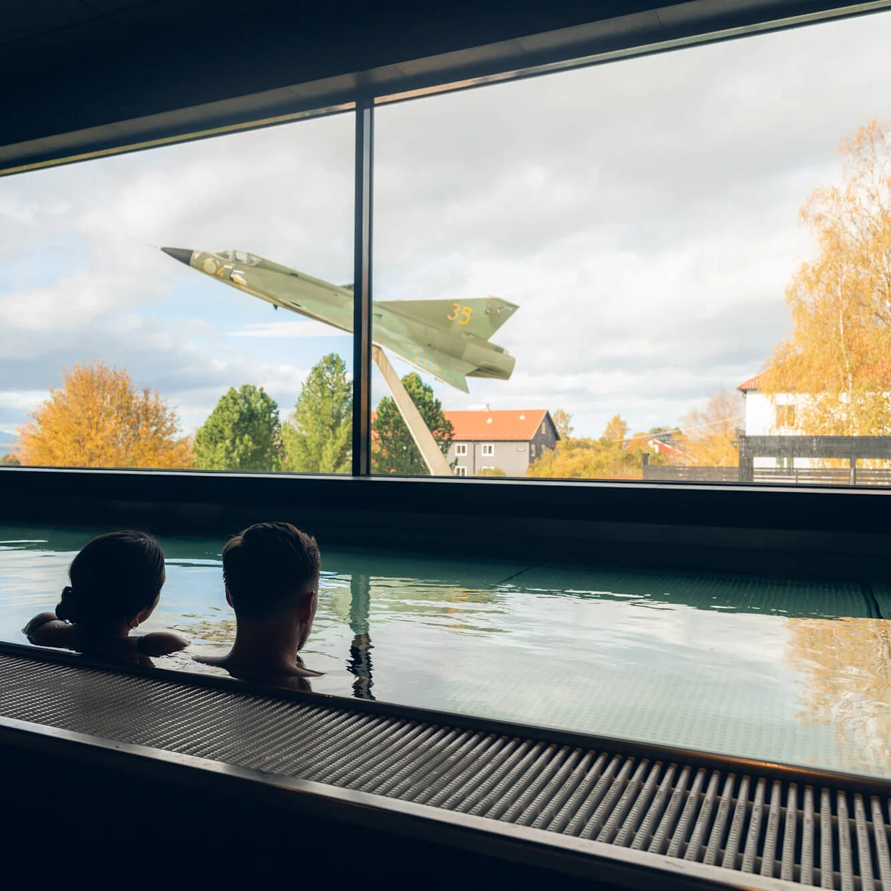 Två personer kopplar av i en inomhuspool, och tittar ut genom ett stort fönster som visar ett monterat stridsflyg och höstträd under en molnig himmel.