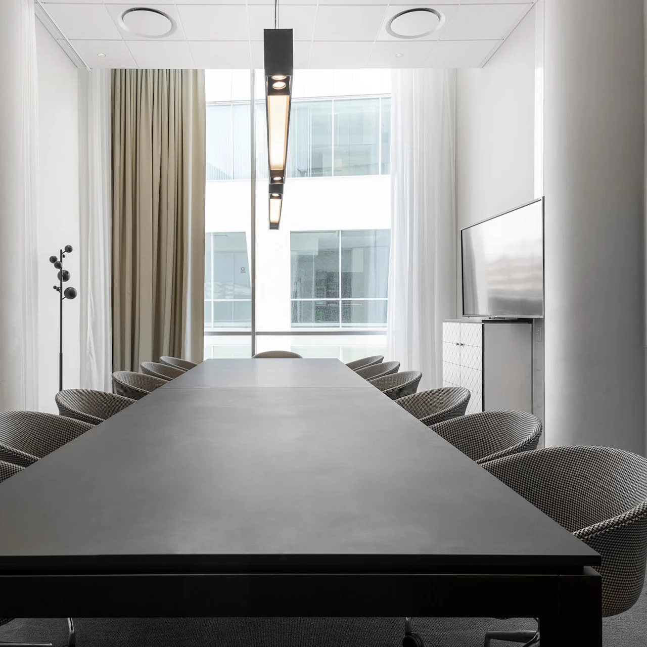 A long black conference table is surrounded by chairs, positioned in a modern office meeting room with large windows, curtains, a television, and overhead lighting.