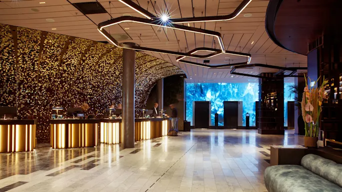Reception area featuring illuminated counters where people interact, surrounded by a golden mosaic wall. Modern lighting hangs overhead, with elevators and blue-lit wall art in the background.