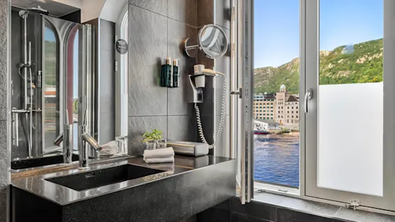 Et moderne badeværelse har en elegant sort håndvask, en vægmonteret hårtørrer og toiletartikler. Der er et vindue med udsigt over vandet og bjergene i det fjerne.