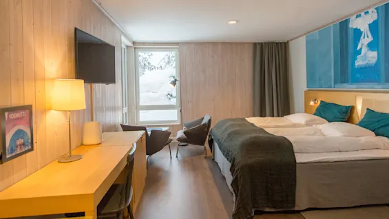 Et hyggeligt hotelværelse har en dobbeltseng, et træbord og stole ved et vindue med udsigt over sneklædte træer. Et vægmonteret TV og en sengelampe giver yderligere bekvemmeligheder. Tekst: "ICEHOTEL".