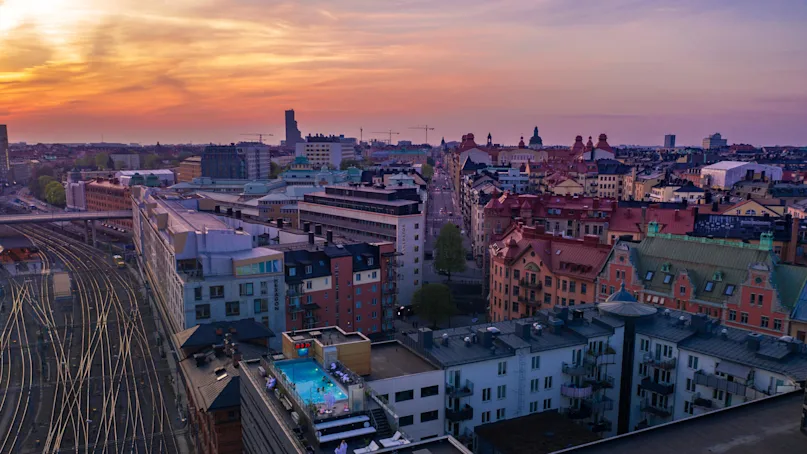 Rooftop swimming pool, surrounded by people, overlooks a sprawling cityscape with sunset-lit buildings and intersecting railway tracks. 