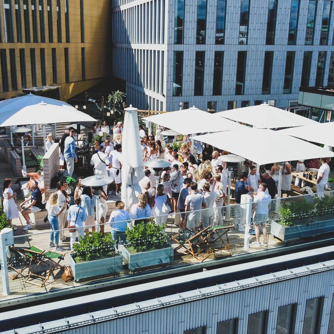 En takterrasse har en samling av folk som sosialiserer under store, hvite parasoller. Omgitt av moderne glassbygg, omfatter scenen dekorative planter og utemøbler.