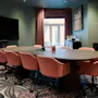 Conference table surrounded by pink chairs, set in a room with a large screen, floral carpet, and artwork. Light filters through windowed curtains, softening the modern decor.