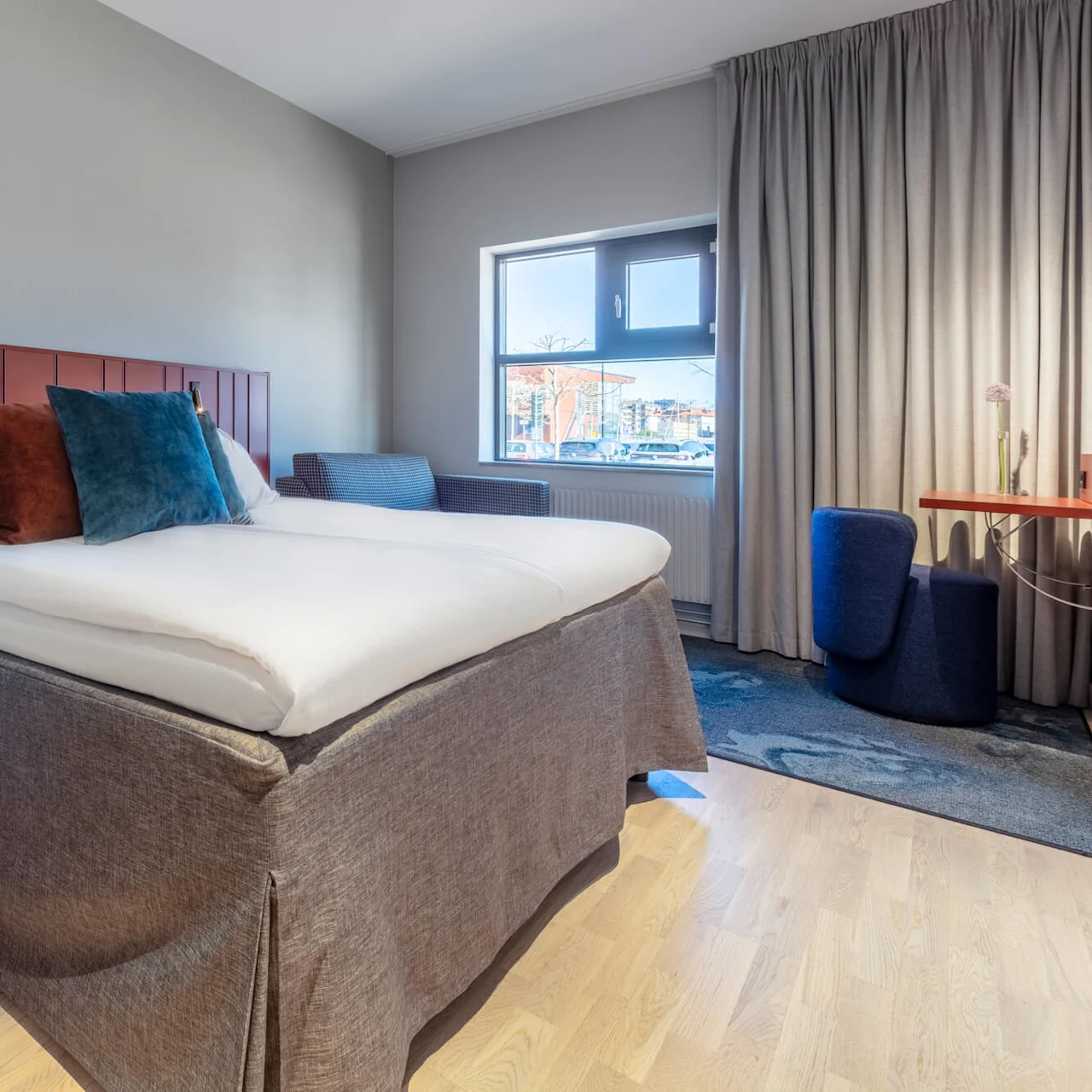 A neatly made bed with pillows against a red headboard, in a bright, modern bedroom with a window and curtains.