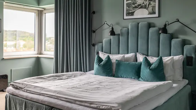 Et komfortabelt hotellsoverom med en plysjseng med turkis sengegavl, mykt sengetøy og en lys vindusutsikt.