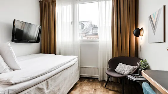 En prydlig säng vilar under en väggmonterad TV i ett litet rum; bruna gardiner inramar ett fönster med stadsutsikt, och en stol med kudde står bredvid en upplyst modern lampa.