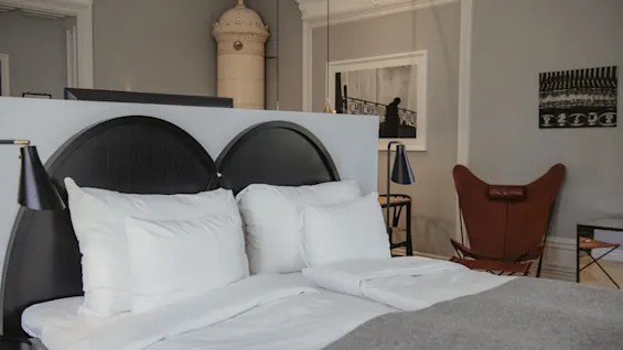 En prydligt bäddad säng med vita lakan och kuddar står i ett stilfullt grått sovrum. En utsmyckad keramisk kamin och modern läderstol tillför elegans till rummet.