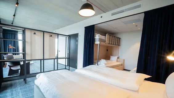 Moderni hotellihuone, jossa on iso sänky ja kerrossänkyalue, erotettu tummalla verhoilla. Pehmeä valaistus valaisee viihtyisän avoimen tilan, jossa on istuimia ja pöytiä.