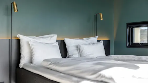 Et moderne hotellrom med en oppredd seng, hvite sengetøy, stilige lamper og et vindu med utsikt.
