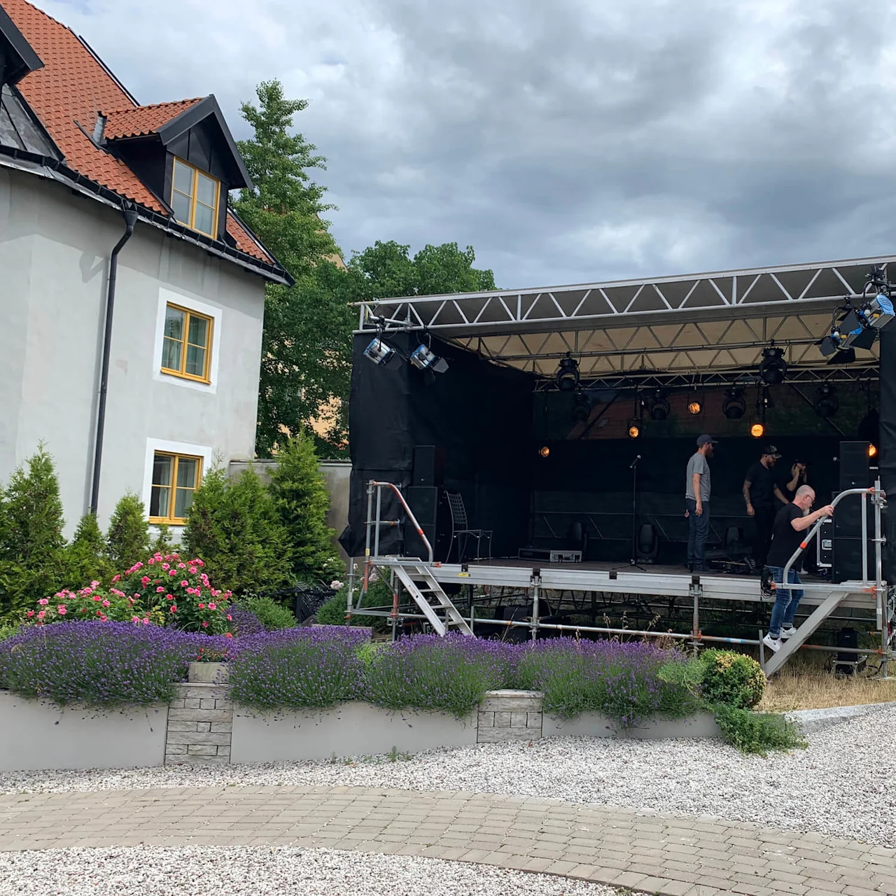 Der opsættes en scene af flere personer i en udendørs have med blomstrende planter og et nærliggende hus. Scenen har ovenlys og er omgivet af grønne områder.