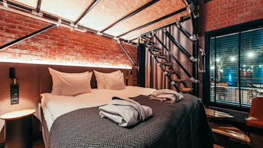 A cozy bed with white pillows and robes is placed against a brick wall. Industrial-style stairs lead upward. Warm lighting provides a comfortable, modern ambiance.