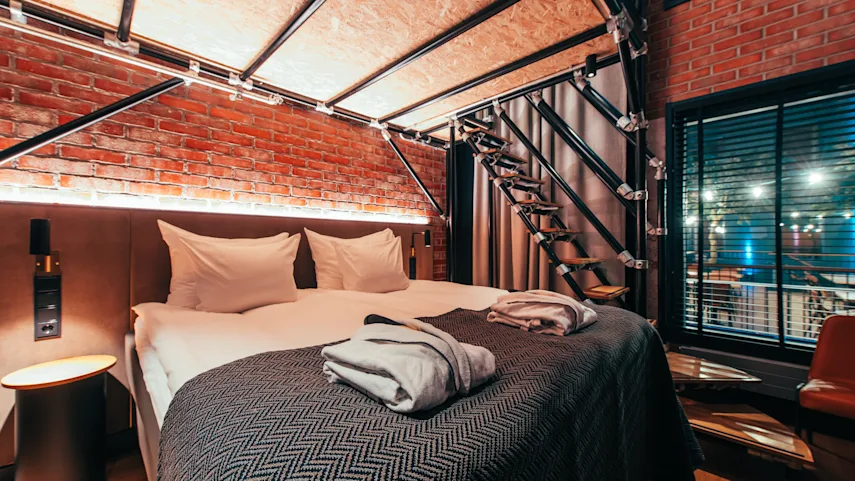 A cozy bed with white pillows and robes is placed against a brick wall. Industrial-style stairs lead upward. Warm lighting provides a comfortable, modern ambiance.
