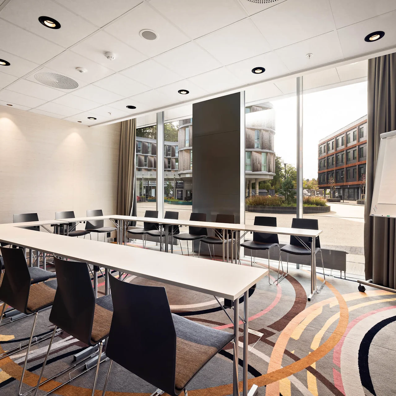 Meeting room with a U-shaped table setup surrounded by black chairs. A blank flipchart and large windows reveal modern buildings outside. Colorful carpet covers the floor.