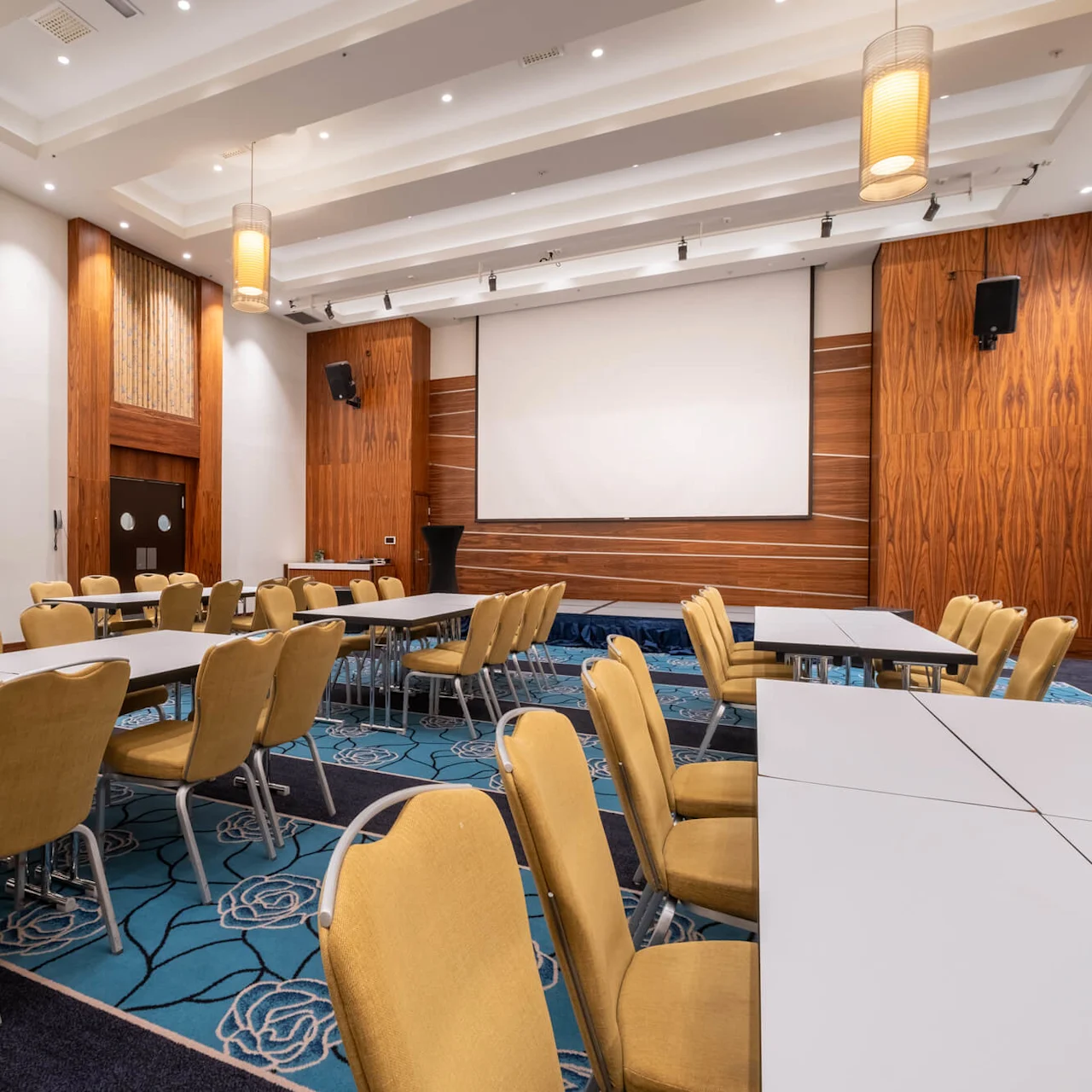 Conference room with empty yellow chairs around long tables, facing a large screen. Wooden panels and floral-patterned blue carpet add to the decor, under warm ceiling lights.