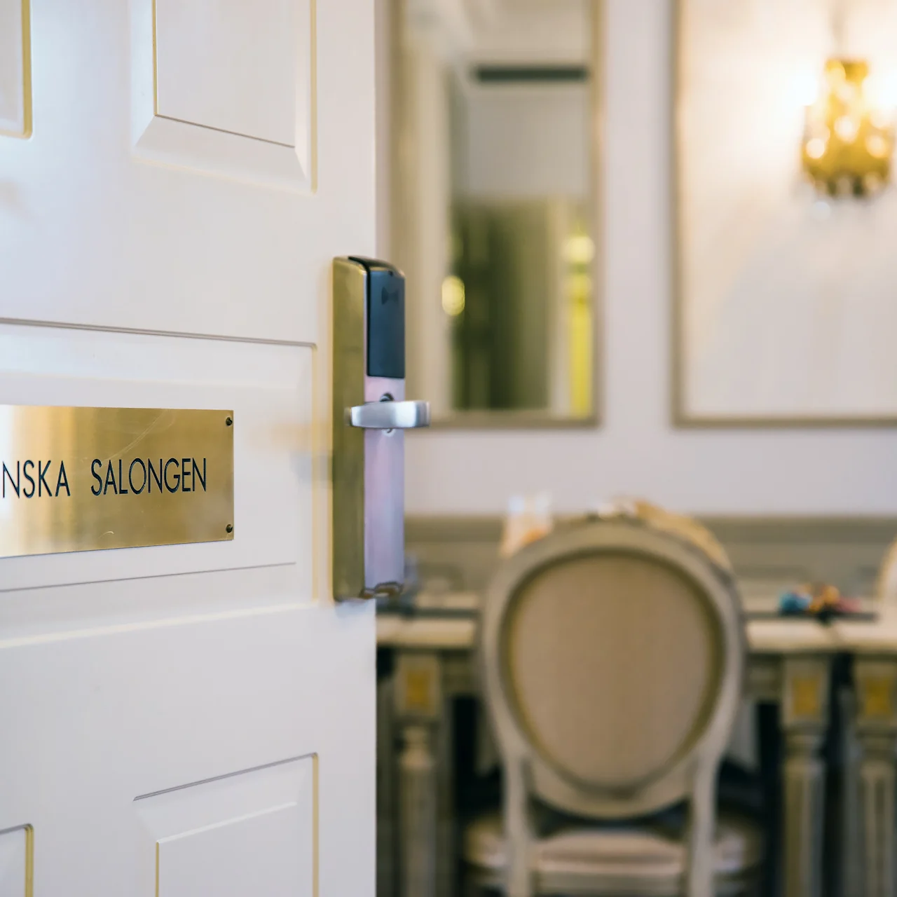 En dörr märkt "FRANSKA SALONGEN" öppnas mot ett elegant rum med utsmyckade stolar runt ett bord, omgiven av inramade speglar och en dekorativ vägglampa.