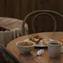 Smörgås med grönsaker och kalkon ligger på en tallrik bredvid en kaffekopp, croissant, skål med flingor och juiceglas. Placeras på ett runt träbord med stolar i en mysig miljö.
