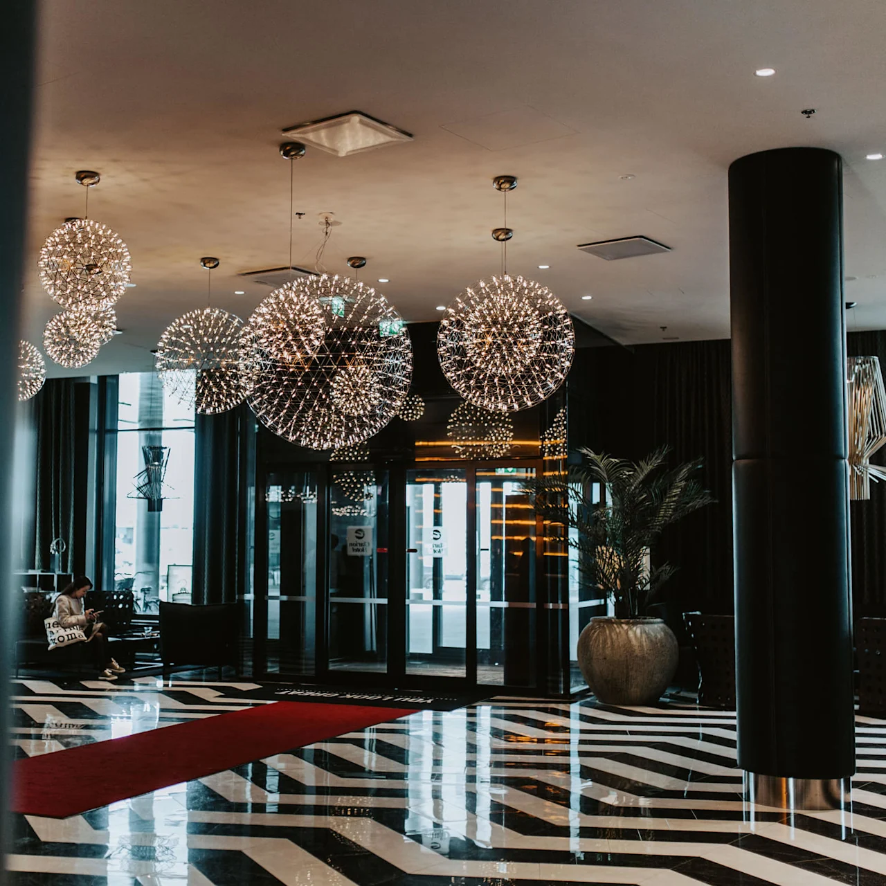 Chandeliers hang above a glossy, patterned floor; a person sits reading in a stylish, modern lobby with large glass doors, potted plants, and a prominent red carpet.