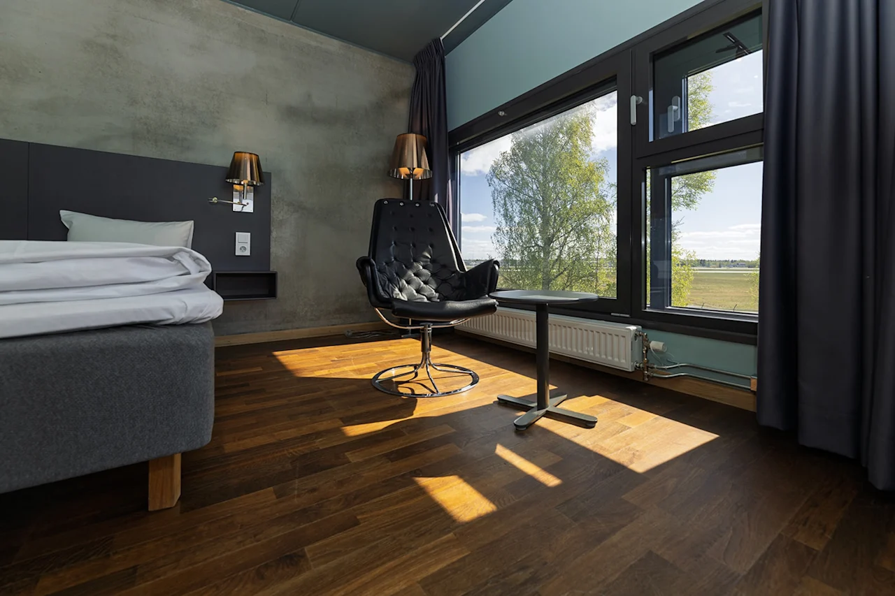 Ett modernt hotellrum med en elegant svart stol som vetter mot ett fönster, bredvid en säng med vita lakan. Solljus strömmar in och lyser upp trägolvet.