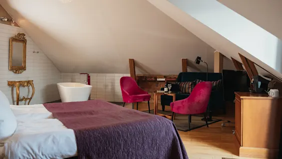 Ett mysigt loftsovrum med en säng med lila filt, ett fristående badkar, rosa stolar och en soffa under sluttande tak, belyst av en ljuskrona.