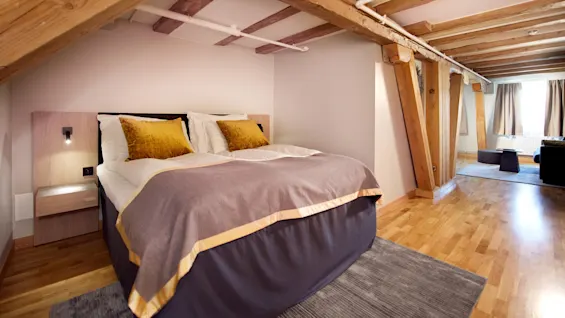 Et lyst soverom med en stor seng, eksponerte trebjelker og moderne møbler, som reflekterer en koselig interiørdesign.