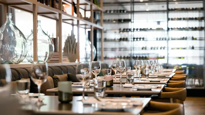 Dining tables, set with glassware and cutlery, are arranged in a sophisticated restaurant. Shelves with large glass bottles and a wine display create an elegant ambiance.