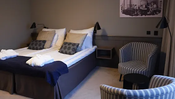 A neatly made twin bed with towels, flanked by bedside tables and lamps, in a subdued hotel room with a striped armchair and a vintage photo on the wall.