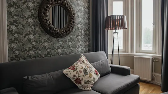 Grå sofa med blomstret pude står mod mønstret væg, dekoreret med en udsmykket cirkulær spejl. En høj lampe oplyser stuen, indrammet af store vinduer.
