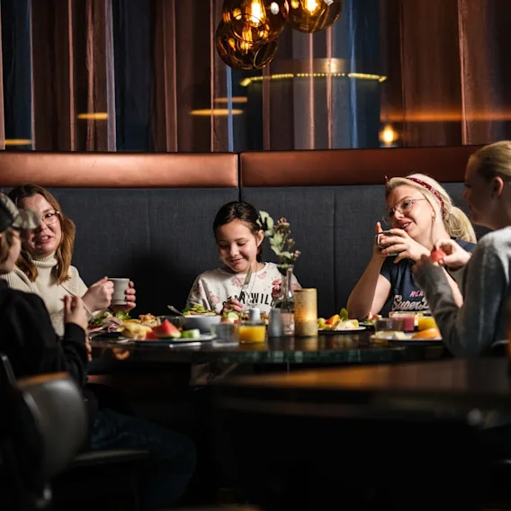 En familie som sitter i en koselig spisesofa rundt et rundt restaurantbord og koser seg med mat.