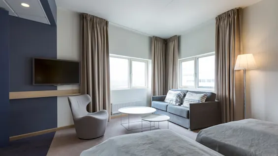 A modern hotel room features twin beds, a gray chair, a wall-mounted TV, a sofa with striped cushions, two round tables, surrounded by beige curtains and soft lighting.