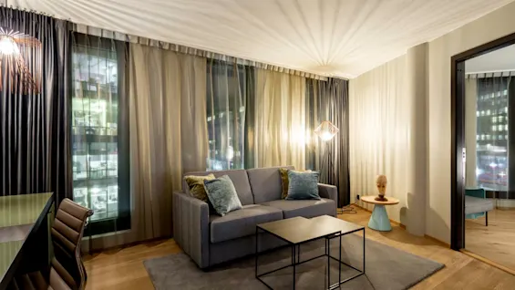 A gray sofa with blue cushions stands on a rug, facing a black coffee table. Soft lighting and floor-to-ceiling curtains create a cozy, modern living room atmosphere.