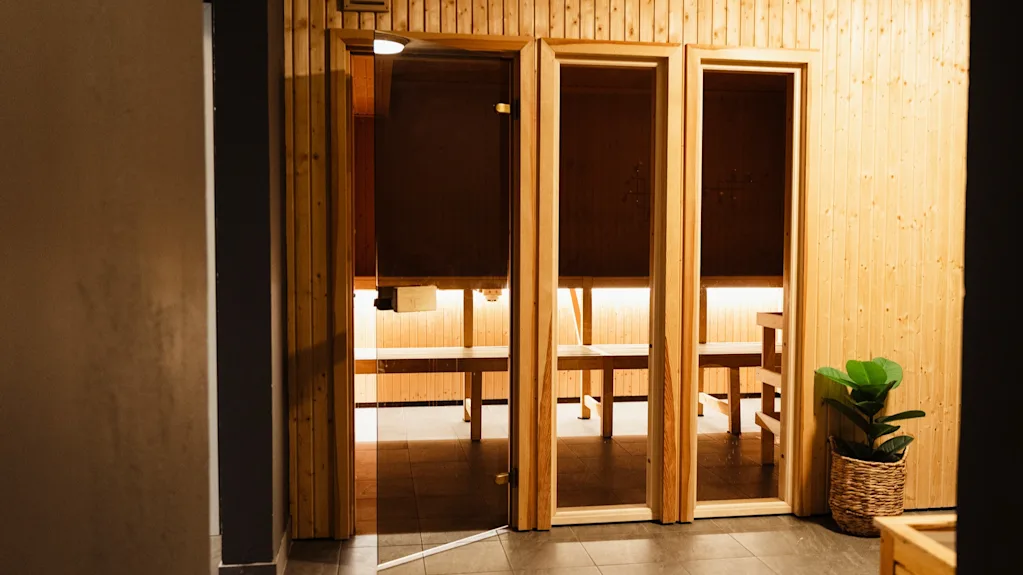 En trädörr till en bastu står på glänt och avslöjar en varmt upplyst interiör med bänkar. En krukväxt står i närheten på ett kaklat golv.