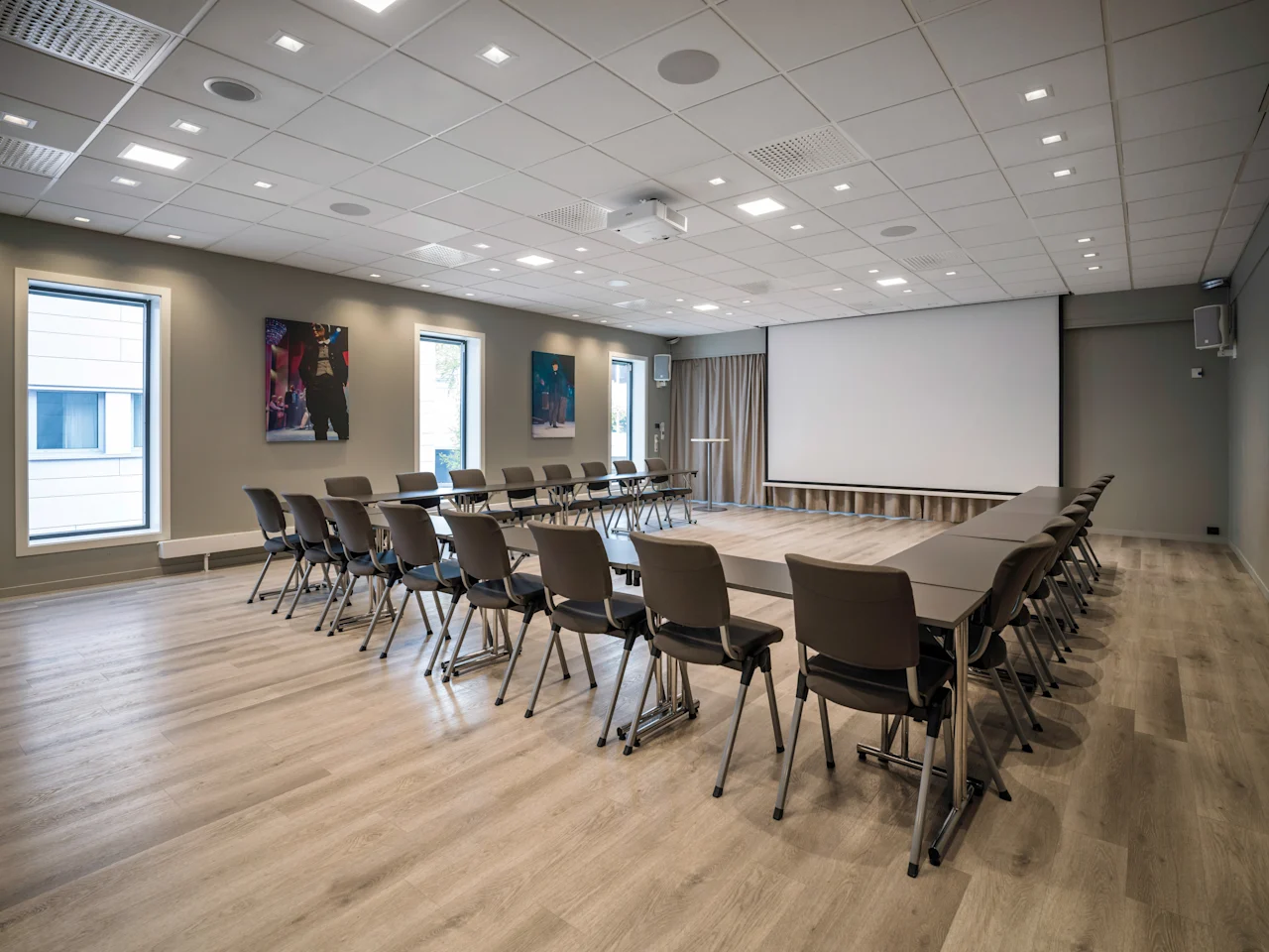 A modern conference room with a U-shaped table setup, chairs, a large projection screen, and windows, ready for a meeting.