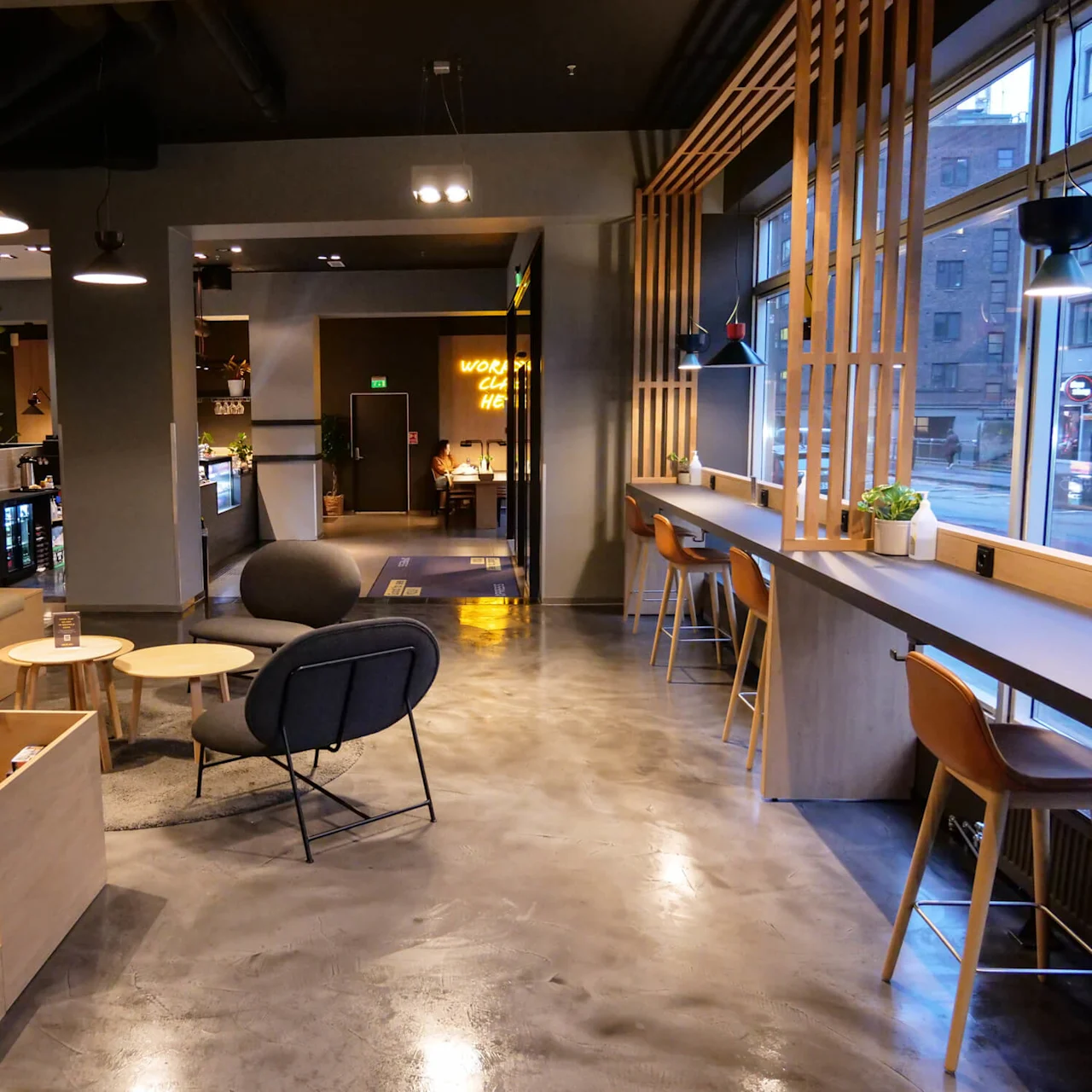 A modern cafe interior with wooden furniture and potted plants. High chairs line the window bar, and cushioned seating surrounds small round tables. The sign reads "WORK CHILL HERE."